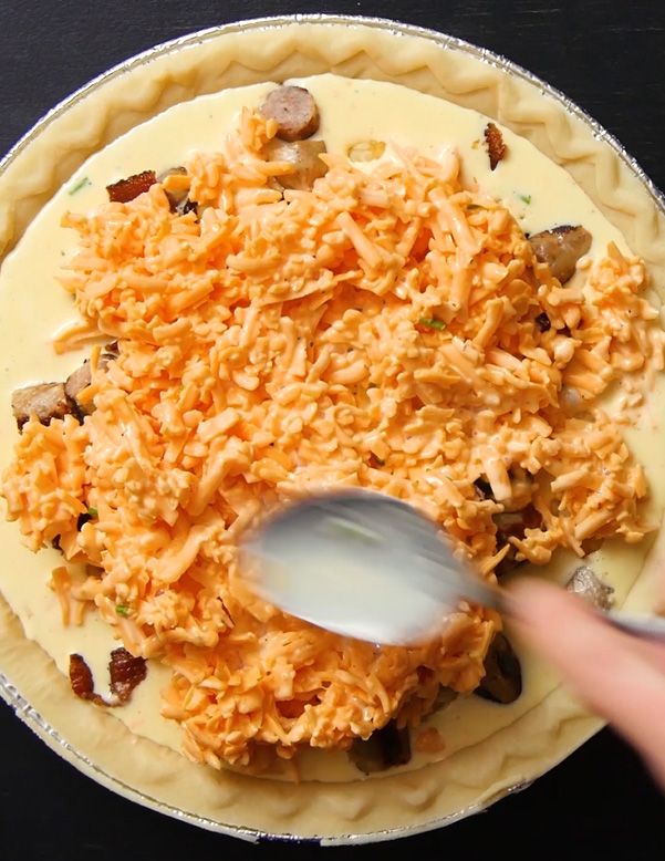 a mountain of cheddar cheese being spread over the top of an uncooked quiche.