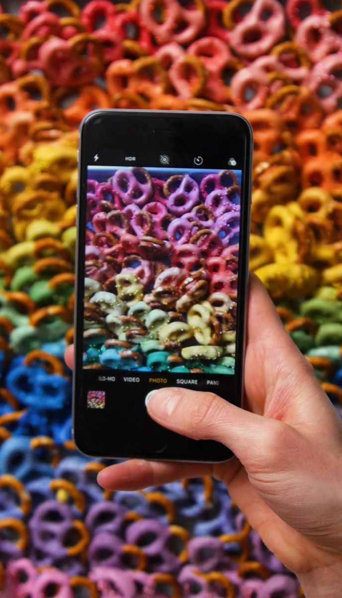 Image may contain an iphone snapping a photo of a cookie tray of rainbow colored pretzels 